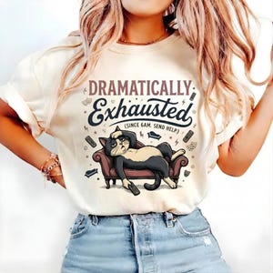 Könnte beinhalten: Cremefarbenes T-Shirt mit dem Text "Dramatisch Erschöpft" und einer Grafik einer schwarz-weißen Katze, die auf einer Couch liegt und eine Fernbedienung hält. Der Text darunter lautet "(Seit 6 Uhr morgens. Hilfe senden)".