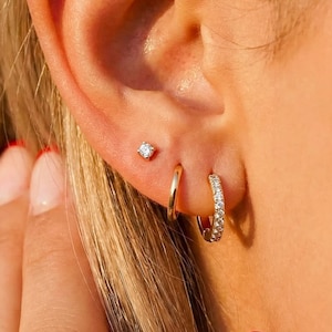 May include: An ear with three earrings: a small stud, a plain gold hoop, and a smaller hoop with gemstones. The ear is next to blonde hair and a hand with red-tipped nails. The earrings are gold in colour. The image is well lit.