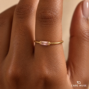 May include: A thin gold band ring featuring a single, horizontally set, rectangular baguette-cut light pink gemstone. The metal band is smooth and polished, providing a simple and clean aesthetic for the jewellery piece worn on a finger.