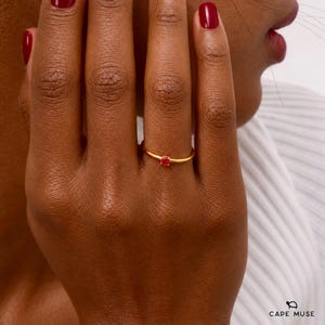 May include: A thin gold band ring featuring a single, small, round, vibrant red gemstone set in a simple prong setting. The ring is worn on a finger, showcasing the polished metallic finish of the band and the bright colour of the stone.