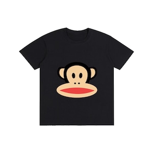 Puede incluir: Camiseta negra con la cara de un mono de dibujos animados. El mono tiene la cara beige, orejas negras y boca roja. La cara del mono está centrada en la parte delantera de la camiseta. La camiseta es de material suave.
