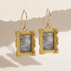 Puede incluir: Un par de pendientes dorados con marcos rectangulares que presentan una pintura en miniatura de una escena de nenúfares. Los marcos tienen detalles ornamentados y están suspendidos de ganchos dorados. Los pendientes se exhiben sobre un fondo neutro.