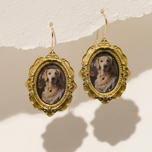 Puede incluir: Un par de pendientes colgantes dorados. Cada pendiente presenta un retrato ovalado de un perro golden retriever dentro de un marco decorativo y ornamentado. El perro lleva un collar. Los pendientes cuelgan de un gancho simple.