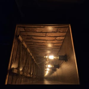 Puede incluir: Un espejo rectangular que refleja un espacio en forma de túnel con ladrillos a la vista, tablones de madera y cuerdas colgantes. El túnel está iluminado por varias bombillas de estilo vintage, creando un ambiente cálido y acogedor. El tema general es rústico e industrial.