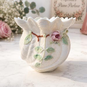 Könnte beinhalten: Eine cremefarbene Keramikvase in Form einer gewellten Muschel mit gerüschtem Rand. Die Außenseite zeigt ein erhabenes, handbemaltes Design einer einzelnen rosa Rosenknospe mit grünen Blättern und einer dünnen, strukturierten braunen Ranke.