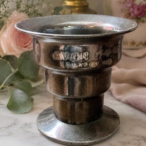 May include: A metallic, tiered pedestal cup with a wide, flared rim and a circular base. The exterior surface features a tarnished silver-tone finish with the embossed text AVON and OUMPEST visible on the upper section of the vessel body.