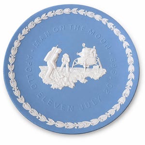 May include: A light blue circular decorative plate featuring white relief detailing. The center depicts two astronauts and a lunar module on the moon's surface. The text reads MAN ON THE MOON APOLLO ELEVEN JULY 20 1969, framed by a white laurel leaf border.