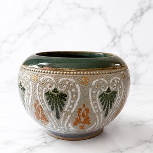 May include: A round ceramic bowl with a dark green glazed rim and a textured light grey body. The exterior features a repeating pattern of white, dark green, and orange floral-like motifs with small gold-coloured accents and a beaded border.