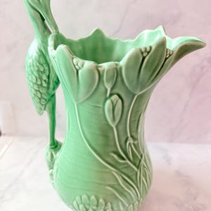 Könnte beinhalten: Ein hellgrüner Keramikkrug mit einem geformten Storchenhenkel und geprägten Blumendetails. Der Körper des Gefäßes zeigt erhabene Tulpenmotive und einen gewellten Rand, wobei die Storchenfigur an der Seite den Henkel des Krugs bildet.