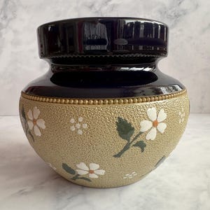 May include: A ceramic vase with a glossy dark blue neck and a textured, light tan body. The lower section features a repeating pattern of white five-petalled flowers with orange centres and dark green leaves, separated from the neck by a beaded rim.