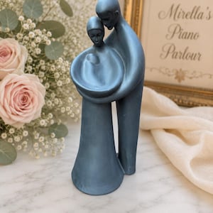 Op de afbeelding: Een glad, matblauw beeldhouwwerk dat twee figuren voorstelt die samenstaan, waarbij één figuur een kleinere vorm in de armen houdt. De figuren hebben langgerekte, gestileerde lichamen en een uniforme, volledig blauwe afwerking.