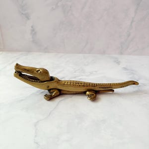 May include: A brass nutcracker shaped like an alligator with a hinged jaw mechanism. The metal figure features a textured back, four short legs, and an elongated tail, with a metallic gold finish and visible joints at the head and body.