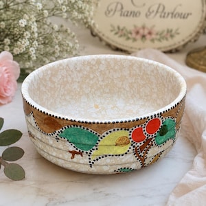 Könnte beinhalten: Eine runde Keramikschale mit gesprenkelter cremefarbener Oberfläche. Die Außenseite zeigt ein handgemaltes Design aus grünen Blättern, gelben Akzenten und roten Beeren, das von einem dekorativen Rand aus kleinen schwarzen Punkten umgeben ist.