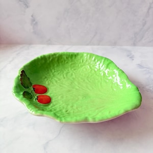 Puede incluir: Un plato de cerámica de color verde brillante con forma de hoja de lechuga texturizada. El plato tiene un acabado brillante y un diseño pintado en el lado izquierdo que muestra dos tomates rojos pequeños con hojas verde oscuro en un tallo marrón.