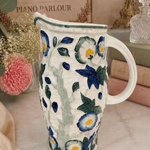 May include: A white ceramic pitcher featuring a textured surface with hand-painted blue and yellow flowers, green leaves, and light green vines. The pitcher has a curved handle, a wide pouring spout, and a decorative rim around the top edge.