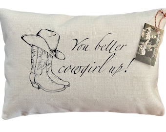May include: A white pillow with the text "You better cowgirl up!" in black. A drawing of a cowboy hat and boots is above the text.