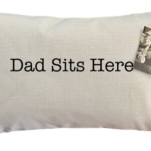 May include: A white pillow with the text "Dad Sits Here" in black lettering. The pillow has a small tag attached to it.