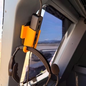 May include: An orange 3D-printed headset holder mounted on a gray surface, holding a black device and a headset. The headset has a microphone and is connected to the black device. The background shows a window with a view of an airport.