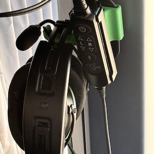 May include: Black gaming headset with a built-in microphone and a control panel. The control panel has buttons and symbols. A green holder is attached to the headset. The headset is connected to a black cable.