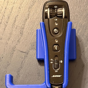 May include: A black Bose headset control module with volume and power buttons, held in a blue 3D-printed holder. The headset control module has a Bluetooth switch and the Bose logo. The holder is mounted on a gray surface.