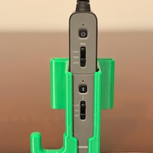 May include: A gray audio device with a black cable is held in a bright green 3D-printed holder. The device has buttons labeled "ANC", "HIGH", "MED", "LOW", "MUTE", "MIX", and "OFF". The brand name "BOSE" is visible.