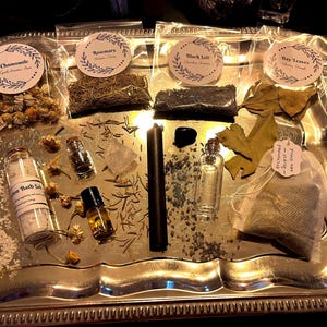 May include: A silver tray holds various items for spiritual practices. Chamomile, rosemary, black salt, and bay leaves are in clear bags with labels. There are also small bottles, a black candle, and a tea bag.