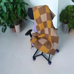 Op de afbeelding: Een gebreide deken met een dambordpatroon in bruin-, beige- en geeltinten is over een zwarte bureaustoel met paarse accenten gedrapeerd. De stoel staat op een witte vloer, met potplanten op de achtergrond.