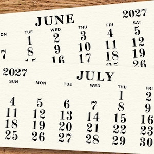 May include: Two cream-colored 2027 calendars, one for June and one for July, are displayed on a wooden surface. The calendars feature black text with the days of the week and dates.