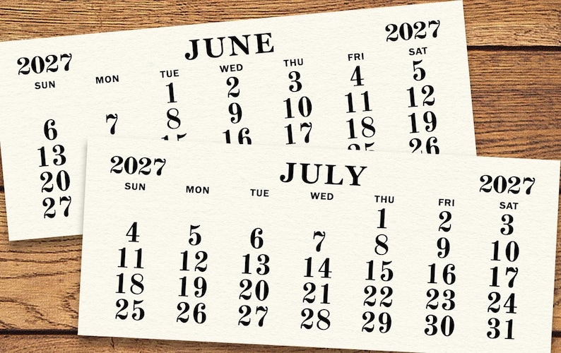May include: Two cream-colored 2027 calendars, one for June and one for July, are displayed on a wooden surface. The calendars feature black text with the days of the week and dates.