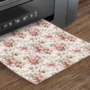 K&ouml;nnte beinhalten: Ein schwarzer Drucker druckt ein Blatt Papier mit einem floralen Muster. Das Muster zeigt rosa Rosen, Schleifen und gr&uuml;ne Bl&auml;tter auf cremefarbenem Hintergrund. Das Papier liegt auf einer Holzoberfl&auml;che.