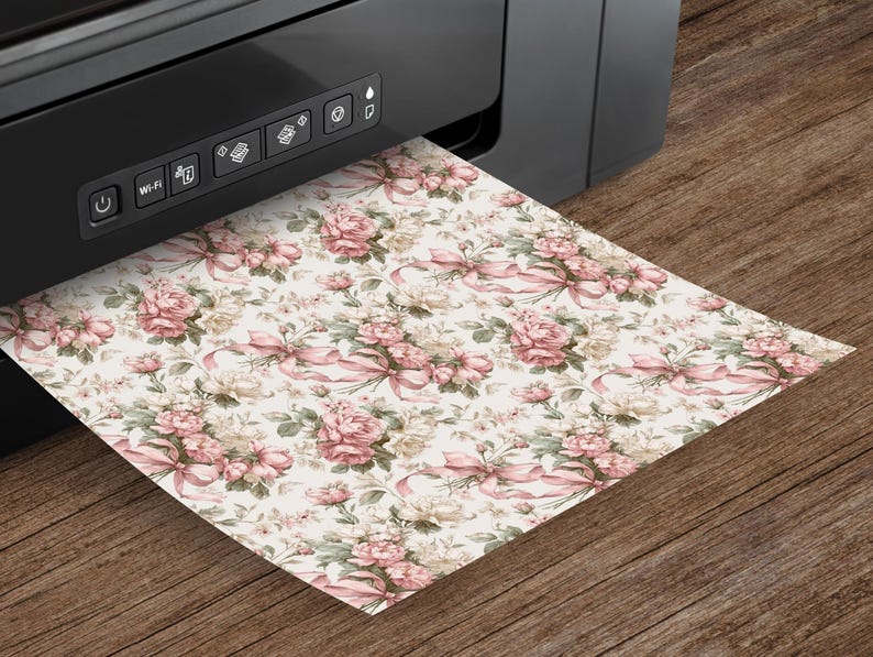 K&ouml;nnte beinhalten: Ein schwarzer Drucker druckt ein Blatt Papier mit einem floralen Muster. Das Muster zeigt rosa Rosen, Schleifen und gr&uuml;ne Bl&auml;tter auf cremefarbenem Hintergrund. Das Papier liegt auf einer Holzoberfl&auml;che.