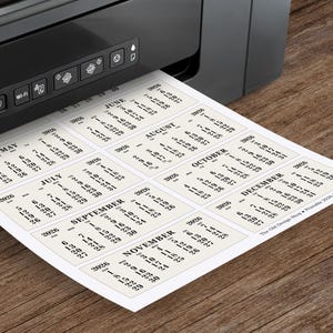 May include: A black printer printing a sheet of paper with calendar cards for the year 2026. Each card displays a month with dates. The paper is cream-colored, and the text is black. The printer is on a wooden surface.