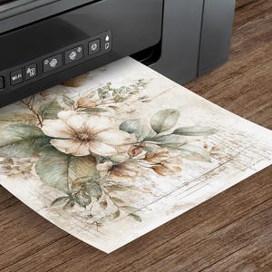 May include: A black printer is printing a watercolor floral design on paper. The artwork features large, cream-colored flowers, green leaves, and smaller brown buds. The paper has a vintage, aged appearance with faint script in the background.