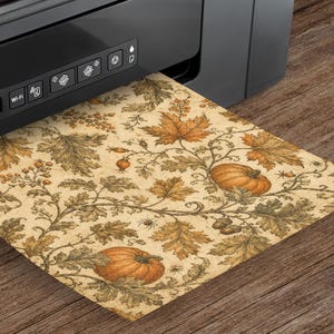 K&ouml;nnte beinhalten: Ein schwarzer Drucker, der ein Blatt Papier mit einem botanischen Muster im Vintage-Stil ausgibt. Das Design zeigt orangefarbene K&uuml;rbisse, braune Eicheln und detaillierte Herbstbl&auml;tter auf beigem Grund, erg&auml;nzt durch kleine Spinnen.
