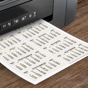 May include: A black printer printing a sheet of calendar cards for 2021. Each card displays a month with dates. The paper is cream-colored, and the text is black. The printer is on a wooden surface.