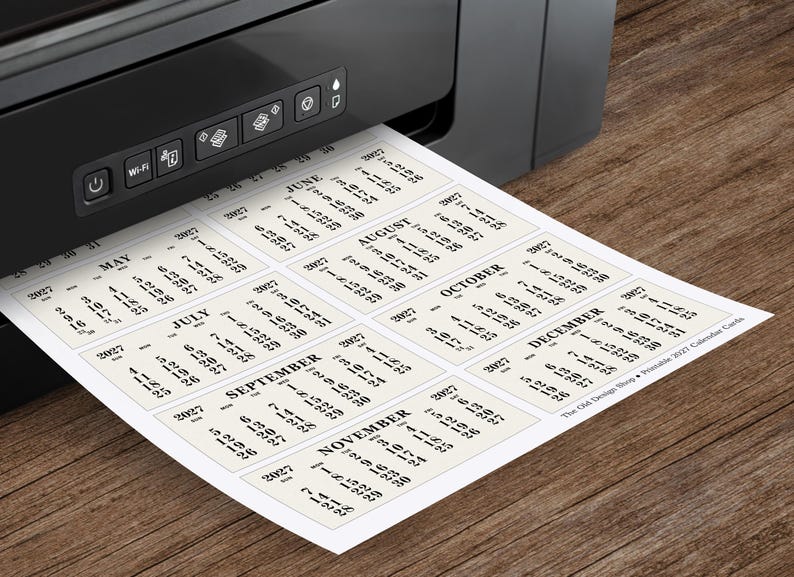 May include: A black printer printing a sheet of calendar cards for 2021. Each card displays a month with dates. The paper is cream-colored, and the text is black. The printer is on a wooden surface.