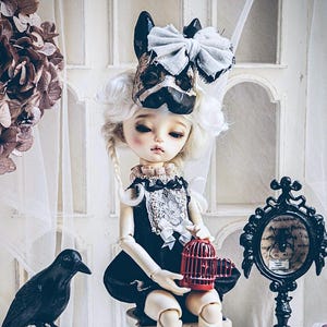 May include: A gothic-themed doll with a dog-faced mask and a large bow sits on a small chair, holding a red birdcage. A black raven sits atop a skull, and a decorative mirror is nearby.