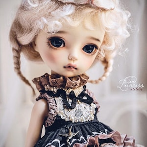 May include: A detailed doll with large, dark eyes and blonde, curly hair. The doll wears a black and brown dress with lace and pearl accents, and a cat-shaped mask rests on its head. The doll's face is delicately painted, and the overall aesthetic is soft and elegant.