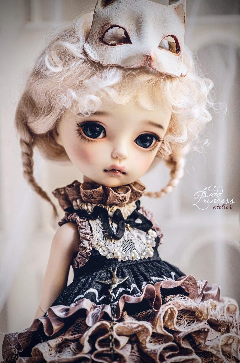 May include: A detailed doll with large, dark eyes and blonde, curly hair. The doll wears a black and brown dress with lace and pearl accents, and a cat-shaped mask rests on its head. The doll's face is delicately painted, and the overall aesthetic is soft and elegant.