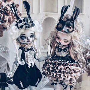 May include: Two gothic-style dolls with elaborate costumes and masks. One doll wears a black and white checkered bunny ear mask, the other a black mask. Both dolls have pale skin, dark eye makeup, and are holding hands. The dolls are surrounded by dried flowers.