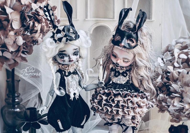 May include: Two gothic-style dolls with elaborate costumes and masks. One doll wears a black and white checkered bunny ear mask, the other a black mask. Both dolls have pale skin, dark eye makeup, and are holding hands. The dolls are surrounded by dried flowers.