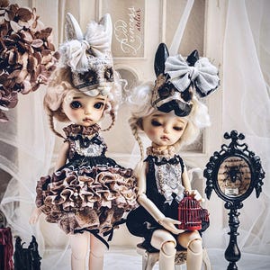 May include: Two porcelain dolls with elaborate costumes and headwear. One doll wears a ruffled skirt and the other sits with a small red birdcage. The dolls have pale skin, blonde wigs, and are set against a soft, blurred background.
