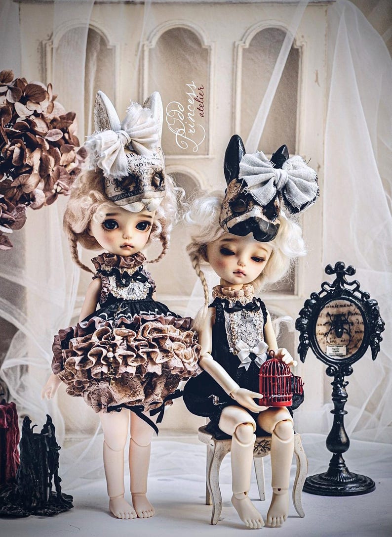 May include: Two porcelain dolls with elaborate costumes and headwear. One doll wears a ruffled skirt and the other sits with a small red birdcage. The dolls have pale skin, blonde wigs, and are set against a soft, blurred background.