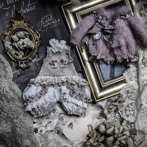 May include: A delicate, light blue and white lace dress with a ruffled hem and a small, white lace bow on the bodice. The dress is displayed on a lace background with a vintage book and a framed photograph.
