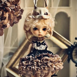 May include: A doll with blonde curly hair and a white rabbit mask. The doll wears a black and tan ruffled dress with a lace collar. The doll has large, dark eyes and a delicate face. The background includes a decorative frame and dried flowers.