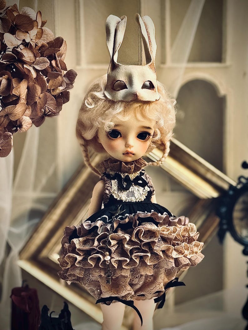May include: A doll with blonde curly hair and a white rabbit mask. The doll wears a black and tan ruffled dress with a lace collar. The doll has large, dark eyes and a delicate face. The background includes a decorative frame and dried flowers.