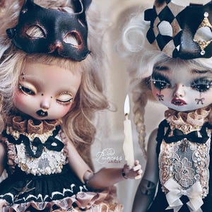 May include: Two art dolls with elaborate makeup and costumes. One doll wears a black cat mask, the other a harlequin-style headpiece. Both dolls have detailed facial features, including dark lipstick and decorative tears. One doll holds a lit candle. The dolls are dressed in black and cream outfits.