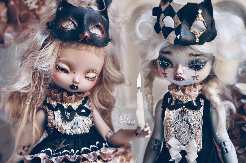 May include: Two art dolls with elaborate makeup and costumes. One doll wears a black cat mask, the other a harlequin-style headpiece. Both dolls have detailed facial features, including dark lipstick and decorative tears. One doll holds a lit candle. The dolls are dressed in black and cream outfits.