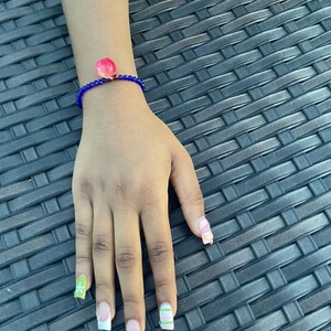 May include: A blue beaded bracelet with a pink flower charm is worn on a hand with painted nails. The nails are painted with various designs, including French tips and colorful stripes. The bracelet is the focal point, set against a textured background.