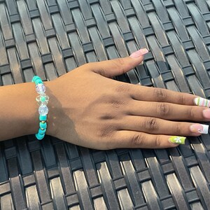 May include: A beaded bracelet with turquoise, clear, and gold beads. The bracelet includes clear beads and a flower-shaped charm. The bracelet is worn on a wrist with manicured nails.